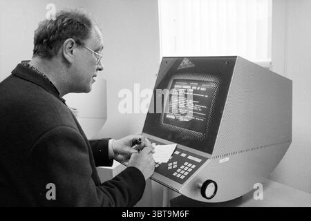 Teun Stam unemployed HBO student, Whizgle News, Dutch Desk, The Netherlands, 1950 - 2000 on 17-03-1994. The image shows these topics. In a softly lit room, a man stands in profile, engaged in a moment of concentration. He wears glasses and has neatly combed hair, dressed in a dark blazer that lends him a slightly formal appearance. In front of him is an unusual, retro-styled computer terminal, its shell showcasing a silver metallic finish with a textured pattern. The screen displays glowing text in bright letters against a darker background, creating a stark contrast that draws the eye. Below Stock Photo