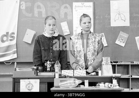 Speech CARA Eenhoornschool, Whizgle News, Dutch Desk, The Netherlands, 1950 - 2000 on 10-02-1991. These topics appear in the image. In a classroom setting, two girls stand side by side in front of a dark chalkboard, which is partly filled with handwritten notes and drawings. The girls are facing forward, exuding a mix of confidence and apprehension. One girl wears a denim jacket over a light-colored shirt, while the other sports a colorful vest layered over a long-sleeve shirt. Both have their hair styled in simple yet neat fashion, with the first girl sporting medium-length hair tied back and Stock Photo