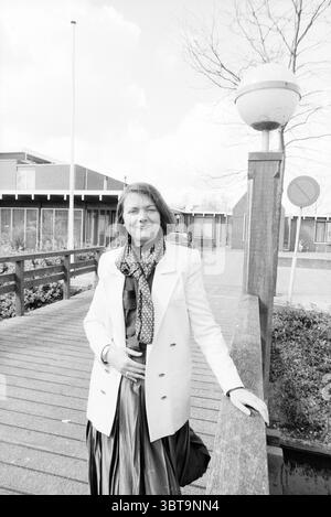 Mrs. de Vrey Mayor Nieuwkoop Mayors Nieuwkoop, Whizgle News, Dutch Desk, The Netherlands, 1950 - 2000 on 11-04-1990. These are the topics in the image. The scene depicts a woman standing confidently on a wooden bridge that stretches over a small body of water. She is dressed in a stylish, light-colored coat that contrasts subtly with the darker tones of her dress, which has a flowing, elegant design. A patterned scarf adds a touch of visual interest and texture around her neck, enhancing her sophisticated appearance. The background features modern buildings characterized by horizontal lines an Stock Photo