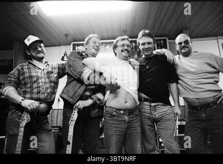 The Bellies Blues Rock Group, Whizgle News, Dutch Desk, The Netherlands, 1950 - 2000 on 01-01-1991. These are the topics in the image. The scene captures a lively gathering of five men standing closely together, exuding a playful camaraderie. Each individual appears to be in high spirits, with broad smiles and casual postures. They are dressed in relaxed, everyday clothing; some wear flannel shirts and jeans, while others have on t-shirts and denim jackets. A couple of them sport sunglasses, adding a touch of coolness to the scene. The setting is an indoor space with warm wooden paneling, givi Stock Photo