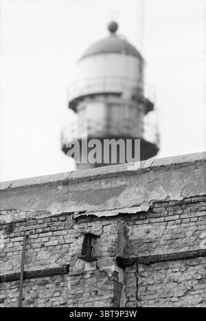 Miscellaneous harbor area, Whizgle News, Dutch Desk, The Netherlands, 1950 - 2000 on 29-03-1993. The image contains these topics. The scene captures a weathered brick wall in the foreground, characterized by its rough texture and visible cracks that suggest age and neglect. Various shades of gray dominate the brick surface, with some areas appearing darker due to shadow play, hinting at a sense of decay. Standing tall in the background is a lighthouse, its rounded tower topped with a distinct dome. The lighthouse features a railing, indicating a viewing platform, and has a utilitarian design t Stock Photo
