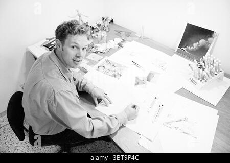 Artist Marco Stoker H'lem Haarlem The Netherlands, Whizgle News, Dutch Desk, The Netherlands, 1950 - 2000 on 26-10-1995. These topics are shown in the image. The scene captures a workspace brimming with creative energy. A man sits at a wooden desk, engaged in drawing or sketching, with various tools spread around him. He wears a light-colored shirt, and his expression is focused yet approachable, suggesting a blend of concentration and enthusiasm. Scattered across the desk are sheets of paper filled with intricate sketches and designs, hinting at an artistic endeavor. Several pens and markers Stock Photo