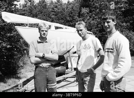 Gebr. Jelles sailors. Sailing, Whizgle News, Dutch Desk, The Netherlands, 1950 - 2000 on 31-07-1986. These topics are shown in the image. In a setting characterized by natural greenery, three young men stand confidently near a sailboat mounted on a trailer. The boat, predominantly white, gleams against the lush backdrop of trees, suggesting a sense of adventure and leisure. The individuals display distinct styles: the first on the left has light blonde hair and is dressed in a blue shirt, paired with dark pants, emanating a casual sporty vibe. The second, with slightly longer blonde hair and a Stock Photo