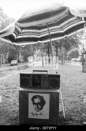 Unveiling of Boman's monument, Whizgle News, Dutch Desk, The Netherlands, 1950 - 2000 on 06-09-1995. These are the topics in the image. In the scene, a vibrant, striped umbrella provides shelter over a vintage boombox placed on a grass patch. The umbrella, adorned with bold patterns of red, black, and white, stands out against a backdrop of muted, overcast skies. Raindrops visibly dot the surface, enhancing the somber, reflective mood that envelops the entire setting. The boombox, featuring a classic boxy design, sits atop a simple, solid-colored pedestal covered partially with a light fabric. Stock Photo
