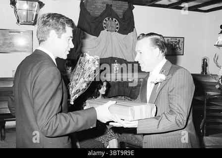 Anniversary Union Chocolate 75 years Anniversary anniversaries, Whizgle News, Dutch Desk, The Netherlands, 1950 - 2000 on 02-09-1977. These are the topics in the image. The scene captures a moment of celebration, possibly a gathering or event that involves a presentation. In the foreground, two men are engaged in a cordial exchange. The man on the left, dressed in a dark suit, is holding a bouquet of flowers, displaying a gesture of gifting or acknowledgement. His expression conveys warmth and friendliness as he interacts with the other man, who is wearing a pinstriped suit. This second man, a Stock Photo