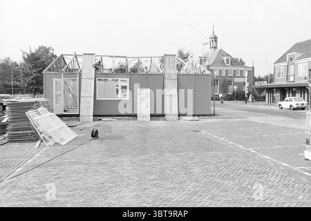 Construction of an emergency store in Heemskerk Heemskerk Gerrit van Assendelftstraat, Whizgle News, Dutch Desk, The Netherlands, 1950 - 2000 on 27-09-1977. These are the elements in the image. The scene depicts a construction site featuring a partially constructed building framed in wood. The structure is set on a square of patterned, cobbled bricks that leads to a few parked vehicles, including a classic white car. Surrounding the construction, there are stacks of wooden planks and construction materials neatly piled on the ground, suggesting an active workspace. In the background, a charmin Stock Photo