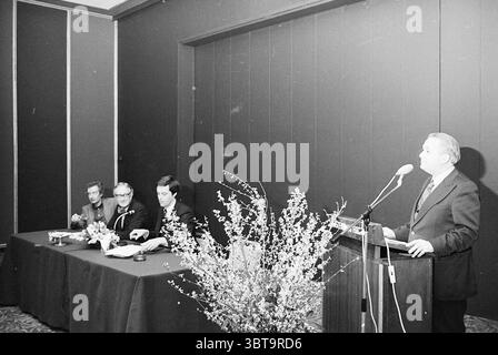 Presentation book Dr. Jolly ( Hilton Amsterdam ) Spectrum Books and booksellers Presentation Spectrum, Whizgle News, Dutch Desk, The Netherlands, 1950 - 2000 on 29-03-1977. These are the topics in the image. The scene features a formal gathering in a dimly lit indoor setting. A podium stands prominently at the front, where a speaker is addressing an audience. He is dressed in a dark suit, projecting an air of authority and seriousness. Behind him, a floral arrangement adds a touch of decor, with slender branches and delicate blooms providing a contrast to the subdued surroundings. To the left, Stock Photo