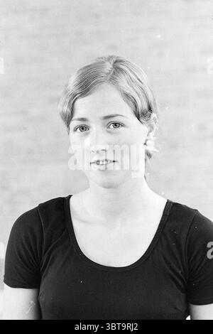 Portraits women's team championships gymnastics, Whizgle News, Dutch Desk, The Netherlands, 1950 - 2000 on 16-07-1970. These are the topics in the image. The scene features a young woman with a soft expression, positioned centrally in the composition. She has light brown hair styled simply, with a few strands lightly framing her face. Her attire consists of a fitted black top, which contrasts with her fair skin. The background presents a smooth, light-toned surface, possibly a wall, which provides a neutral backdrop that emphasizes her features. The lighting is soft and even, casting a gentle Stock Photo