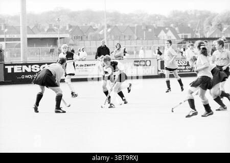 Alliance - Keep Fit Hockey, Whizgle News, Dutch Desk, The Netherlands, 1950 - 2000 on 13-11-1988. These topics are shown in the image. In a dynamic outdoor scene, two teams engage in an intense field hockey match on a well-maintained pitch. A group of players, clad in coordinated uniforms, moves energetically across the surface. The athletes primarily wear dark shorts and light tops, contrasting against the solid color of the field. One player, positioned centrally, grips the hockey stick with both hands, poised to make a decisive play. He appears focused and determined, while several opponent Stock Photo