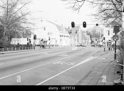 Exterior of Restaurant De Geleerde Man Bennebroek Rijksstraatweg, Whizgle News, Dutch Desk, The Netherlands, 1950 - 2000 on 02-1989. The image includes these topics. The scene captures a quiet, urban intersection, imbued with a tranquil mood. The foreground features a wide road, its surface marked with faint lines indicating lanes and a dedicated bus lane. On one side of the road, a bicycle path runs parallel, edged by a low wooden fence, hinting at an environmentally conscious city. Across the intersection, a variety of buildings create an eclectic architecture, characterized by both modern a Stock Photo
