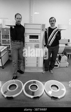 2 system administrators Chamber of Commerce Chamber of Commerce and factories, Whizgle News, Dutch Desk, The Netherlands, 1950 - 2000 on 17-01-1990. The image includes these topics. In the setting, two men stand prominently, each holding large, circular data storage reels in a well-lit room filled with computing equipment. The first man, on the left, wears a fitted black shirt and light blue jeans, showcasing a modern style of the era. He has a slight smile and stands confidently, with his body angled slightly towards the viewer. The second man on the right dons a striped pullover that contras Stock Photo