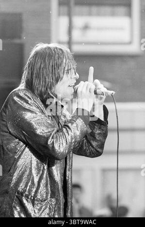 Liberation pop butter market Music Haarlem Botermarkt The Netherlands, Whizgle News, Dutch Desk, The Netherlands, 1950 - 2000 on 05-05-1987. These are the elements in the image. The scene features a captivating performance under a dimly-lit atmosphere, where a male vocalist takes center stage. He is dressed in a shiny, sequined jacket that catches the light with every movement, enhancing the energy of the moment. The jacket is predominantly black with flecks of silver and reflects light dramatically, creating a contrast against the darker elements of the background. The vocalist's hair is long Stock Photo