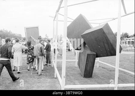 Unveiling statue of Sancta Maria Noordwijkerhout Revelations Noordwijkerhout, Whizgle News, Dutch Desk, The Netherlands, 1950 - 2000 on 26-06-1984. These are the topics in the image. The scene features a lively outdoor gathering centered around a striking abstract sculpture. The sculpture consists of large, interlocking geometric forms—some cubes and flat rectangular shapes—positioned at various angles, creating a sense of dynamic tension. The artwork is framed by a simple, white wooden structure, emphasizing its bold geometry. Surrounding the sculpture, a diverse group of people engages with Stock Photo