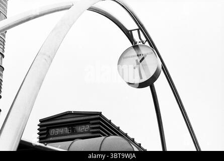 Millennium clock, Whizgle News, Dutch Desk, The Netherlands, 1950 - 2000 on 23-11-1998. These are the elements in the image. The scene is dominated by an architectural structure characterized by sleek, modern lines and a metallic finish. A prominent, large circular clock hangs from an elongated, curved arm that arches gracefully above. The clock's face features a minimalist design, with clear distinctions between the hours and a simplistic layout, all presented in monochromatic tones. Below the clock, a dark-colored base sits, likely housing electronic elements. It has a structured, layered ap Stock Photo