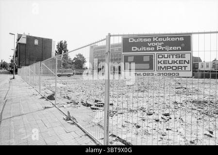 Signs 'German Kitchen Import' attached to construction fence along vacant lot Spaarndamseweg corner of Transvaalstraat Haarlem Spaarndamseweg The Netherlands, Whizgle News, Dutch Desk, The Netherlands, 1950 - 2000 on 29-07-1991. The image includes these topics. The scene features an empty lot, marked by a rugged terrain of exposed earth and scattered debris, suggesting recent demolition or construction. A chain-link fence surrounds the area, offering an unobstructed view of the interior while providing a stark separation from the adjacent spaces. In the background, a couple of buildings can be Stock Photo