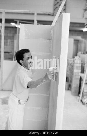 Mason training courses, Whizgle News, Dutch Desk, The Netherlands, 1950 - 2000 on 21-05-1995. The image includes these topics. In a bustling indoor construction setting, a man is seen applying a smooth layer of plaster to a partially constructed wall. He is wearing a light-colored shirt, which contrasts with the dust and mist of plaster residue that clings to his clothes. His expression reflects concentration and focus, indicative of the meticulous nature of his task. The wall he is working on is made of rectangular blocks, and to the left, partially visible, there's a second wall behind him, Stock Photo