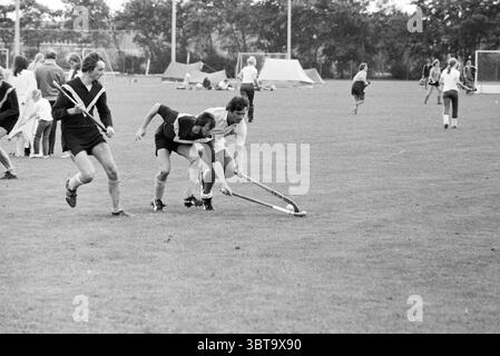 Final hockey tournament Alliance Hockey, Whizgle News, Dutch Desk, The Netherlands, 1950 - 2000 on 24-08-1980. These are the elements in the image. The scene captures a dynamic moment on a large grassy field, where a spirited game is taking place. In the foreground, three individuals are engaged in an intense play, two of them clad in dark uniforms while one wears a light-colored outfit. The player in the center is bending low, skillfully maneuvering a stick that extends towards the ball, creating a sense of urgency and action. Surrounding them, several others populate the field, participating Stock Photo