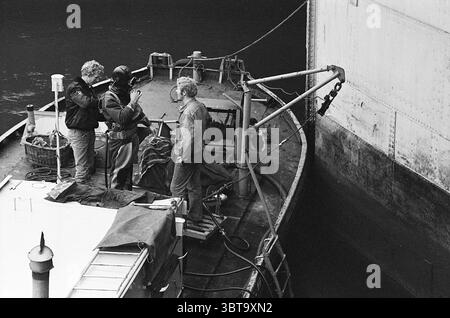 Work at the lock Work, Whizgle News, Dutch Desk, The Netherlands, 1950 - 2000 on 13-10-1969. The image includes these topics. The scene depicts a small boat docked next to a large structure, likely a warehouse or a ship. Three individuals are situated on the deck of the boat, engaged in conversation. One person stands to the left, dressed in a dark jacket, and seems focused on the interaction with the others. The figure in the center is wearing a wet suit, suggesting they may be involved in underwater work or diving. The third individual, positioned on the right, has lighter hair and wears a d Stock Photo