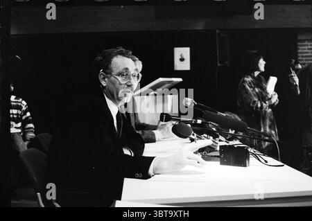 Press conference H'dorp train disaster Hoofddorp The Netherlands, Whizgle News, Dutch Desk, The Netherlands, 1950 - 2000 on 29-11-1991. The image contains these topics. The scene captures a formal setting, likely a press conference or announcement. In the foreground, a man sits at a sleek, white table, dressed in a suit and tie, exuding a serious demeanor. He has glasses perched on his nose, and his expression is one of focus as he gestures with his hand, perhaps emphasizing a point. Behind him, two additional figures are partially visible. One appears to be taking notes, while the other, slig Stock Photo