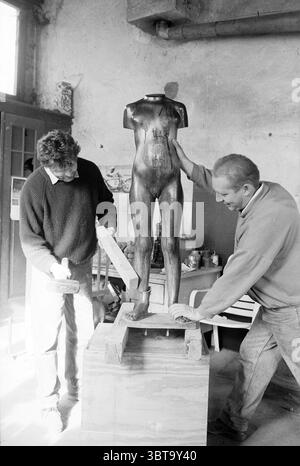 Binder foundry Haarlem The Netherlands, Whizgle News, Dutch Desk, The Netherlands, 1950 - 2000 on 21-10-1992. The image shows these topics. In a rustic workshop filled with a warm, inviting atmosphere, two individuals are collaboratively working on a sculpture. The room has a slightly worn, vintage feel, with textured walls that evoke a sense of history. Sunlight filters in through a large window, casting soft shadows that dance across the floor and illuminate the scene. The central focus is a bronze mannequin, unfinished yet striking, resting on a wooden pedestal. Its smooth, metallic surface Stock Photo