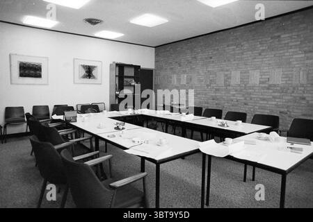 Meeting room containing a wall with old reliefs of a Stations of the Cross ('converted chapel Raphaelstichting Santpoort'), Whizgle News, Dutch Desk, The Netherlands, 1950 - 2000 on 15-02-1985. The image shows these topics. In a neutral-toned room, a spacious rectangular table takes center stage, surrounded by a series of black chairs arranged neatly. The table is covered with white tablecloths, giving it a clean and organized appearance. On the surface of the table, several small white teacups are placed alongside saucers, suggesting an opportunity for refreshments. A few plates and a couple Stock Photo