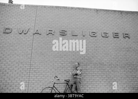 Paul van der Made Dwansligger, Whizgle News, Dutch Desk, The Netherlands, 1950 - 2000 on 05-09-1994. These topics appear in the image. The scene portrays a man standing against a textured, light-colored brick wall that has an industrial feel, characterized by its uniformity and simplicity. The wall features prominent lettering that suggests a name or possibly a title, positioned at the top and slightly to the left. The man, dressed in a casual outfit, leans slightly against the wall, his arms crossed with a relaxed demeanor. He has a gentle expression and appears to be enjoying the moment, pos Stock Photo