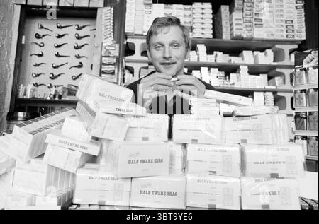 Mr Busbroek with many packs of rolling tobacco cigars and cigarettes, Whizgle News, Dutch Desk, The Netherlands, 1950 - 2000 on 04-05-1983. The image shows these topics. In a quaint, rustic shop filled with a variety of tobacco products, a young man sits amidst a chaotic arrangement of neatly stacked boxes. The boxes, predominantly white with faint designs, are piled high around him, creating a barrier that hints at a sense of organization within the apparent disarray. The young man exudes a friendly demeanor, with a slight smile on his face, suggesting a welcoming atmosphere. He has light hai Stock Photo