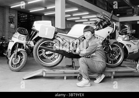 Mechanic with Yamaha motorcycles Motorcycles motorcycle racing, Whizgle News, Dutch Desk, The Netherlands, 1950 - 2000 on 15-07-1985. The image shows these topics. The scene features a person in a sporty, tracksuit-like outfit, crouching beside a sleek motorcycle. The individual has short hair and demonstrates a relaxed yet confident posture, resting their arms on the side of the bike. The motorcycle is predominantly white with vibrant red and black accents, showcasing a bold racing number '1' on its side. Surrounding this centerpiece, several other motorcycles can be seen in the background, c Stock Photo