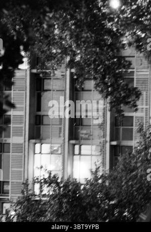 Vacant flats Schalkwijk Haarlem The Netherlands, Whizgle News, Dutch Desk, The Netherlands, 1950 - 2000 on 24-08-1993. These topics appear in the image. The scene is primarily illuminated by a soft, ambient glow emanating from several windows of a modern building, suggesting the presence of activity within. The architecture features a series of rectangular windows framed by sleek lines, with an overall structured and minimalist design. Surrounding the building, there are dark outlines of trees, their leaves creating a natural frame that contrasts with the geometric shapes of the building. The Stock Photo