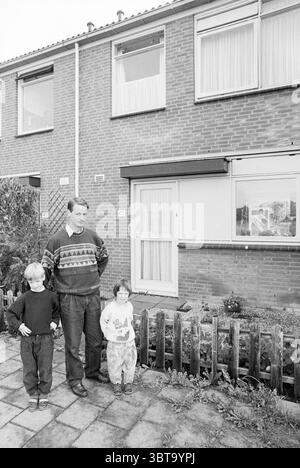 Father with 2 children in front of the house, Whizgle News, Dutch Desk, The Netherlands, 1950 - 2000 on 09-1991. The image contains these topics. In a residential setting, a man stands alongside two children in front of a modest brick house. The building features a simple design, characterized by its rectangular shape and multiple windows through which light streams in. The façade is a warm reddish-brown, complemented by a grassy patch and a low wooden fence that outlines the foreground. The man, appearing to be in his late 30s or early 40s, has short hair and is dressed in a patterned sweater Stock Photo