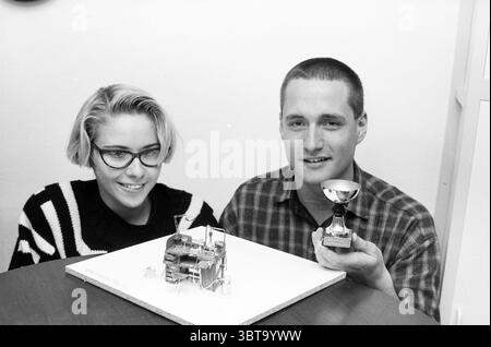 Two people with award-winning miniature workbench, Whizgle News, Dutch Desk, The Netherlands, 1950 - 2000 on 11-1989. The image includes these topics. In a well-lit indoor setting, two individuals sit at a round wooden table. The backdrop is a plain, light-colored wall that provides a stark contrast to the subjects. One person has a short, stylish haircut and wears black-rimmed glasses. They are dressed in a bold, monochromatic striped sweater that adds visual interest. The other person, seated beside them, has a clean-cut appearance and is wearing a checkered shirt in muted tones. On the tabl Stock Photo