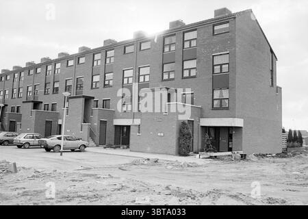 Work on the construction of a new residential area, Whizgle News, Dutch Desk, The Netherlands, 1950 - 2000 on 11-1981. The image shows these topics. The scene captures a row of modern, rectangular residential buildings with a brick facade, suggesting a contemporary architectural style. There are three levels, each featuring multiple windows framed in a minimalist design, allowing for a geometric rhythm across the structure. In the foreground, the ground is unfinished and rugged, indicating ongoing construction or recent development, with patches of dirt and debris scattered throughout. Two par Stock Photo