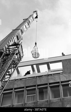 Hoisting satellite globe post office IJm. IJmuiden The Netherlands, Whizgle News, Dutch Desk, The Netherlands, 1950 - 2000 on 27-09-1994. These are the elements in the image. The scene presents a dynamic construction setting, characterized by a large crane towering over a building. The crane, with its robust metallic structure, angles downwards, holding a large, white container suspended from a thick cable. This container appears sturdy and industrial, contrasting sharply against the backdrop. The building, with its solid concrete facade, showcases a blend of gray and beige tones, adding a sen Stock Photo