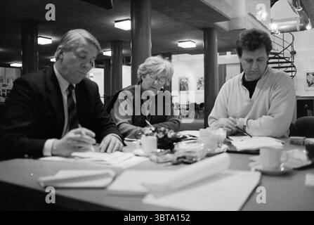 Oogkamp Foundation, Whizgle News, Dutch Desk, The Netherlands, 1950 - 2000 on 14-02-1994. The image shows these topics. In a spacious, modern workspace characterized by minimalist design, three individuals are engaged in a focused discussion around a central table. The table is strewn with various documents, notepads, and pens, suggesting a brainstorming or planning session. To the left, a man dressed in a dark suit intently writes, his brow slightly furrowed as he leans forward, reflecting concentration. His hair is neatly styled, and he appears to be deep in thought, possibly analyzing the m Stock Photo
