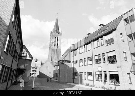 New construction around old church including use of fisheye camera lens, Whizgle News, Dutch Desk, The Netherlands, 1950 - 2000 on 10-1985. The image includes these topics. The scene presents a quaint urban landscape characterized by brick buildings that are primarily rectangular and have large windows. The structures are in varying shades of gray, reflecting a monochromatic palette that offers a sense of calm and nostalgia. In the background, a prominent church tower rises, its intricate design marked by a tall spire crowned with a point. The tower features a series of arches and a clock face Stock Photo