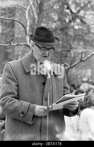 Liberation Day in Haarlem Haarlem The Netherlands, Whizgle News, Dutch Desk, The Netherlands, 1950 - 2000 on 05-05-1980. The image contains these topics. In the scene, an elderly man stands at a microphone, delivering a speech or reading from a paper. He wears a dark, wide-brimmed hat and thick glasses, which rest on his nose. His coat appears to be a heavy, textured fabric, likely gray or a similar muted tone, suggesting a chill in the air. The background is filled with a blurred crowd, indicative of an outdoor gathering, though individuals are not distinctly visible. The people seem engaged, Stock Photo