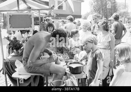Creative market Santpoort South Market Santpoort-Zuid, Whizgle News, Dutch Desk, The Netherlands, 1950 - 2000 on 05-06-1982. These topics appear in the image. In a vibrant outdoor market scene, a variety of people gather around a central figure engaged in pottery. The potter, a man with a muscular build and sun-kissed skin, sits on a wooden stool, carefully shaping clay with his hands. He wears only denim shorts, indicating a warm and sunny day. Around him, a small group of curious children, with wide eyes and eager expressions, lean in closer, captivated by the craft. One child, a boy with to Stock Photo