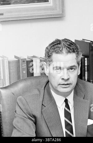 Portrait man., Whizgle News, Dutch Desk, The Netherlands, 1950 - 2000. The image contains these topics. The scene features a middle-aged man seated in an office setting. He has neatly combed hair with a slight wave, and his expression conveys seriousness and focus. He is dressed in a tailored suit, predominantly gray with a subtle sheen, and wears a crisp white shirt complemented by a dark tie featuring diagonal stripes. The background reveals a well-organized bookshelf filled with various volumes, suggesting a scholarly or professional environment. The books are predominantly in muted tones o Stock Photo