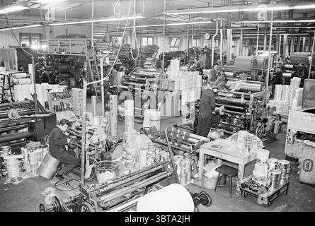 Fa.Ruys packaging material Kleine Houtweg Haarlem interiors of the new and old factory, Whizgle News, Dutch Desk, The Netherlands, 1950 - 2000 on 20-12-1962. The image includes these topics. The scene captures a bustling industrial environment filled with machinery and workers engaged in various tasks. The overall palette leans toward monochrome, with shades of gray dominating the space, which adds a vintage, atmospheric quality. In the foreground, two workers are focused on their tasks. One is seated beside a large machine, carefully inspecting a component, while the other stands, adjusting t Stock Photo