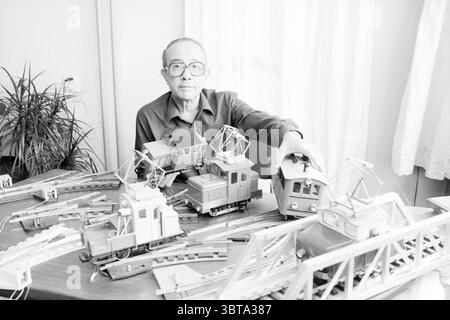 Hans Schoof with trains Hobby Trains, Whizgle News, Dutch Desk, The Netherlands, 1950 - 2000 on 08-10-1987. The image includes these topics. The scene depicts an indoor setting, filled with a warm, nostalgic atmosphere. A soft, diffused light filters in through sheer curtains, casting subtle shadows across the space. In the foreground, a middle-aged man sits at a table cluttered with an assortment of model trains and railway components. He has a contemplative expression, wearing glasses that lend him an air of thoughtfulness. His clothing is simple and understated, reflecting a sense of practi Stock Photo
