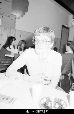 Portrait of a young woman with glasses at the table in a canteen-like room, Whizgle News, Dutch Desk, The Netherlands, 1950 - 2000 on 14-04-1979. The image shows these topics. The scene captures a retro atmosphere, characterized by a distinct vintage aesthetic. A young woman sits at a table, her short hair styled in a chic bob. She wears large, round glasses that accentuate her expressive eyes. Her attire is simple, yet stylish, with a light-colored top. Surrounding her, the background reveals a gathering place filled with people engaged in conversations. Several individuals are visible at oth Stock Photo