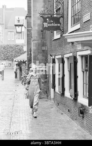 Ingrid Keune for fashion section People, Whizgle News, Dutch Desk, The Netherlands, 1950 - 2000 on 08-06-1988. The image includes these topics. In a charming street scene, a narrow cobblestone pathway winds through a quaint neighborhood. On one side, there are traditional brick buildings with feature windows adorned in white frames; some are slightly ajar. The buildings show a weathered texture, hinting at their age and history. A young person strolls along the path, dressed in a light-colored overall that contrasts softly with the darker tones of the surrounding bricks. They have a cheerful e Stock Photo