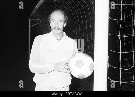 Footballer Fred Bischot ADO'20, Whizgle News, Dutch Desk, The Netherlands, 1950 - 2000 on 01-11-1987. These are the topics in the image. The scene is set on a soccer field at night, illuminated by a few overhead lights that cast a warm glow over the area. A man stands beside a goalpost, creating a focal point in the composition. He has a notable mustache and a slight smile, wearing a light-colored tracksuit that contrasts with the darker ambient surroundings. In his hands, he holds a soccer ball marked with distinct black and white pentagons and hexagons, adding an element of playful contrast Stock Photo