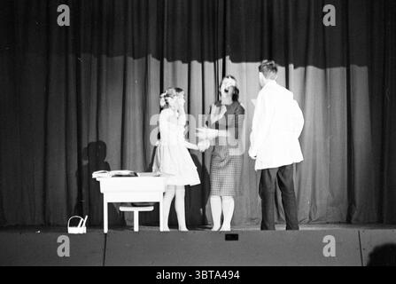 Farewell evening Primary school Germaine de Boer Theater, Whizgle News, Dutch Desk, The Netherlands, 1950 - 2000 on 27-07-1967. These are the topics in the image. On a stage draped with deep, rich curtains, three figures interact in a lively scene. The curtain's folds create a dramatic backdrop, casting gentle shadows that enhance the depth of the setting. In the foreground, two women engage in conversation. One woman, dressed in a light-colored, knee-length dress with delicate detailing, exudes youthful exuberance. Her hair is styled in soft waves, framing her face as she gestures animatedly. Stock Photo
