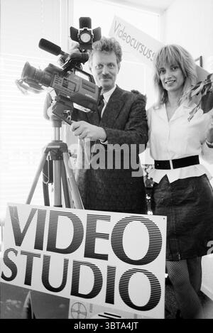 Mr v.d. More video course Zandvoort Courses Video Zandvoort, Whizgle News, Dutch Desk, The Netherlands, 1950 - 2000 on 23-03-1990. These are the topics in the image. The scene captures two individuals positioned prominently in a well-lit studio environment. One person is holding a professional video camera, standing confidently on the left side. They sport a checkered suit that adds a touch of retro charm to their appearance, complemented by a slight smile that conveys enthusiasm. The other person stands on the right, dressed in a stylish outfit featuring a white blouse and a black skirt. Thei Stock Photo