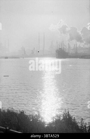 Atmosphere photo North Sea Canal IJmuiden Atmosphere, Whizgle News, Dutch Desk, The Netherlands, 1950 - 2000 on 15-06-1973. These are the elements in the image. The scene presents a hazy industrial landscape, characterized by a calm body of water that reflects a shimmering path of light across its surface. The water appears soft and serene, contrasting with the ruggedness of the surrounding environment. In the distance, silhouettes of towering smokestacks and industrial structures punctuate the skyline, hinting at the busy activities of a harbor or factory. These dark forms fade into a muted b Stock Photo