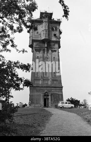 Restoration + exterior water tower IJmuiden. Restoration Water tower IJmuiden The Netherlands, Whizgle News, Dutch Desk, The Netherlands, 1950 - 2000 on 04-05-1981. These are the elements in the image. In the scene, a tall, imposing tower dominates the landscape, showcasing a sturdy, rectangular structure with a clock face prominently displayed near its top. The tower's exterior has an industrial, utilitarian design, characterized by a muted color palette of grays and browns that lend it an austere feel. Surrounding the tower, the ground is grassy, with patches of greenery interspersed, creati Stock Photo