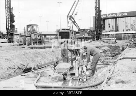 Construction of Schiphol Station Tunnel Parking parking lots parking lots Schiphol Schiphol, Whizgle News, Dutch Desk, The Netherlands, 1950 - 2000 on 27-04-1989. These topics appear in the image. The scene depicts a bustling construction site with a monochrome aesthetic that emphasizes the industrial atmosphere. In the foreground, two workers are engaged in tasks related to the construction process. They are clad in protective clothing, suggesting a focus on safety amid the ongoing work. One worker is leaning over machinery, closely inspecting instruments, while the other appears to be manipu Stock Photo