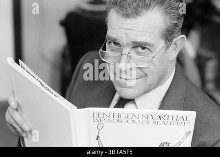 Portrait of Mr Berk KNVB with information folder 'Your own sports hall. Dream or reality', Whizgle News, Dutch Desk, The Netherlands, 1950 - 2000 on 18-11-1986. The image shows these topics. In this scene, an older gentleman is seated, holding a book close to his chest. He is wearing a dark suit, complemented by a light-colored dress shirt, and he sports a slightly playful smirk, suggesting he is in a relaxed or jovial mood. His hair is gray and neatly combed back, and he wears glasses that sit comfortably on his nose, framing his sharp, observant eyes. The background is slightly out of focus, Stock Photo