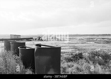 Sand extraction pit Nieuwemeer Sand, Whizgle News, Dutch Desk, The Netherlands, 1950 - 2000 on 25-10-1978. These topics are shown in the image. The scene presents a wide, open landscape that merges land and sky in a gentle gradient. In the foreground, several large, dark containers sit in a row, their curved edges hinting at industrial use. The surface of the containers appears weathered, with patches of rust visible, indicating an age and story behind them. Beyond the containers, a vast expanse of land stretches out, layered with muted shades of gray and brown. The ground is uneven, with vari Stock Photo